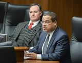 Attorney General Keith Ellison sits at the testifiers' table and speaks to someone out of view. Representative Lesch sits next to him.